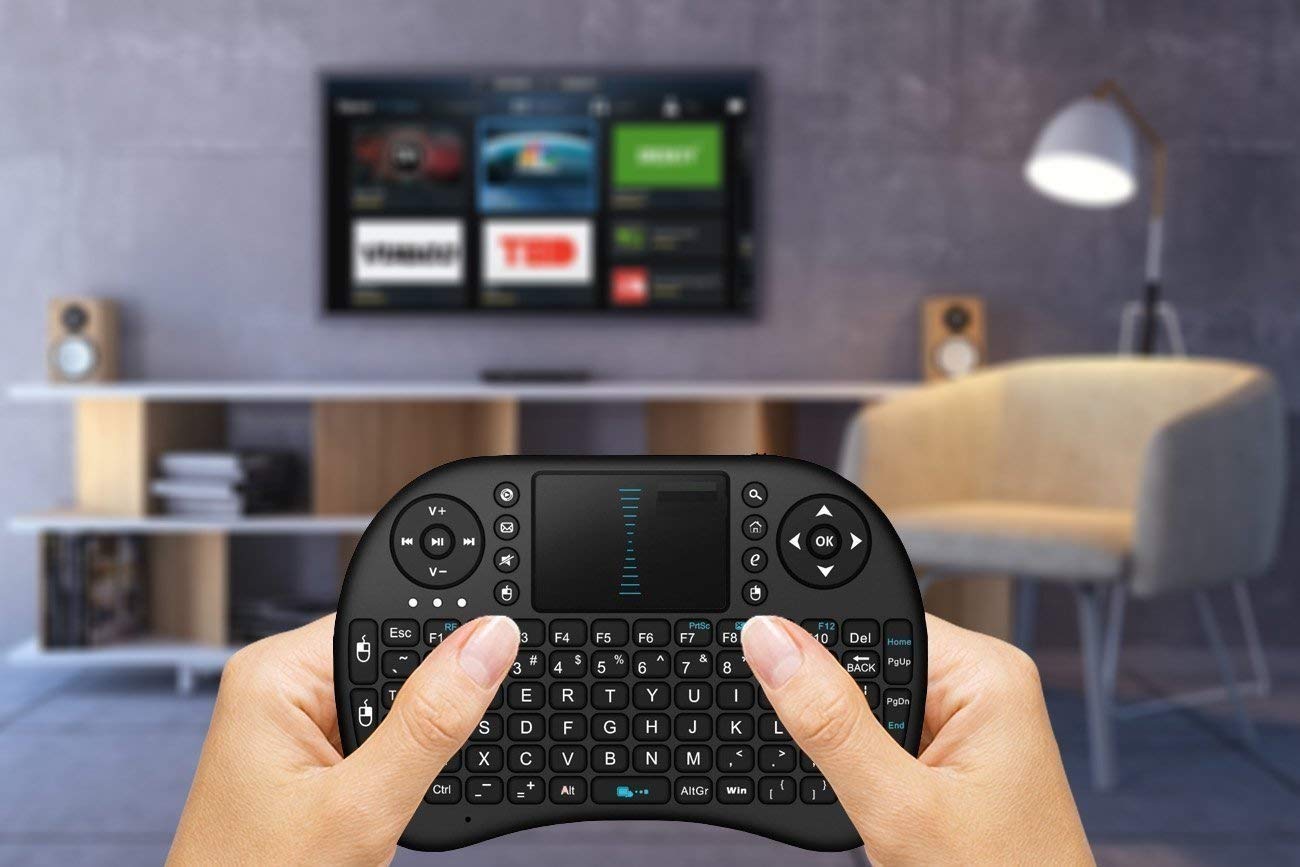 Hand holding a small keyboard remote control in front of a TV in a living room setting.
